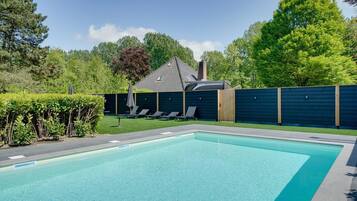 Ein rechteckiger Pool mit Fliesenrand, umgeben von einem dunklen Zaun, und ein Gebäude mit geneigtem Dach im Hintergrund.