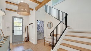 A staircase with wooden steps and black railings, a woven light fixture, a door with a window, and a wall with a circular mirror and decorative wall art. - Sail Away Estate: 5BDR with Private Pool (Tybee Island)