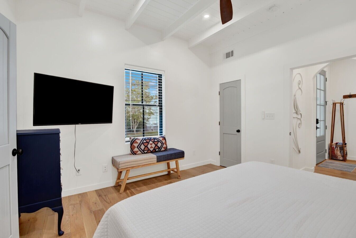 A bedroom with a large bed, a bench with a patterned cushion, a television mounted on the wall, a door, and a window with blinds.