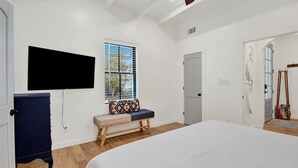 A bedroom with a large bed, a bench with a patterned cushion, a television mounted on the wall, a door, and a window with blinds. - Sail Away Estate: 5BDR with Private Pool (Tybee Island)