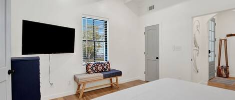 A bedroom with a large bed, a bench with a patterned cushion, a television mounted on the wall, a door, and a window with blinds.