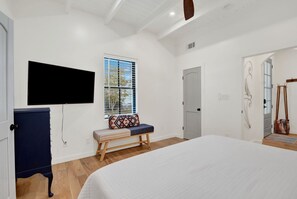Ein Schlafzimmer mit einem großen Bett, einer Sitzbank mit gemustertem Kissen, einem an der Wand befestigten Fernseher, einer Tür und einem Fenster mit Jalousien.