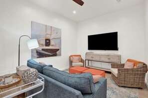 A modern living room with a sofa, ottoman, and a flat-screen TV. - Sail Away Estate: 5BDR with Private Pool (Tybee Island)