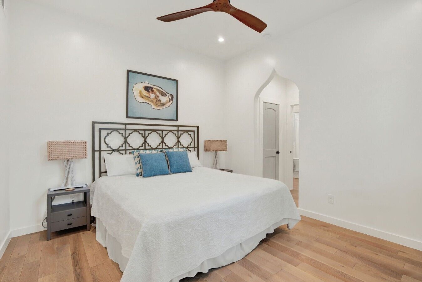 A bedroom with a bed, two bedside tables, a ceiling fan, a framed artwork, and a door leading to another room.