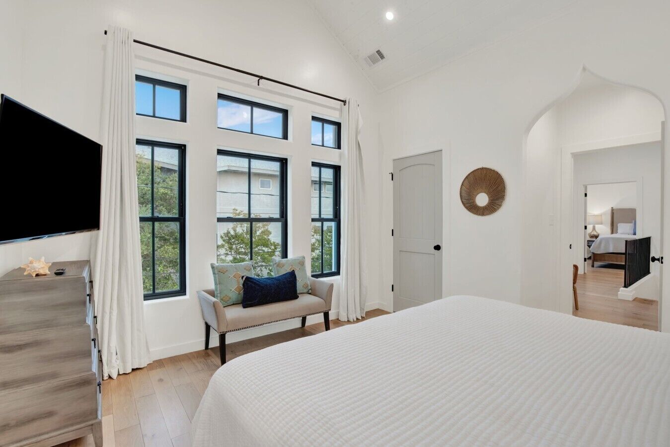A bedroom with a large bed, a sofa, and a TV. There are multiple windows and a door.