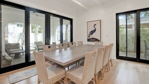 A spacious dining area with a long table and wicker chairs, large sliding glass doors, and a framed bird painting on the wall. - Sail Away Estate: 5BDR with Private Pool (Tybee Island)