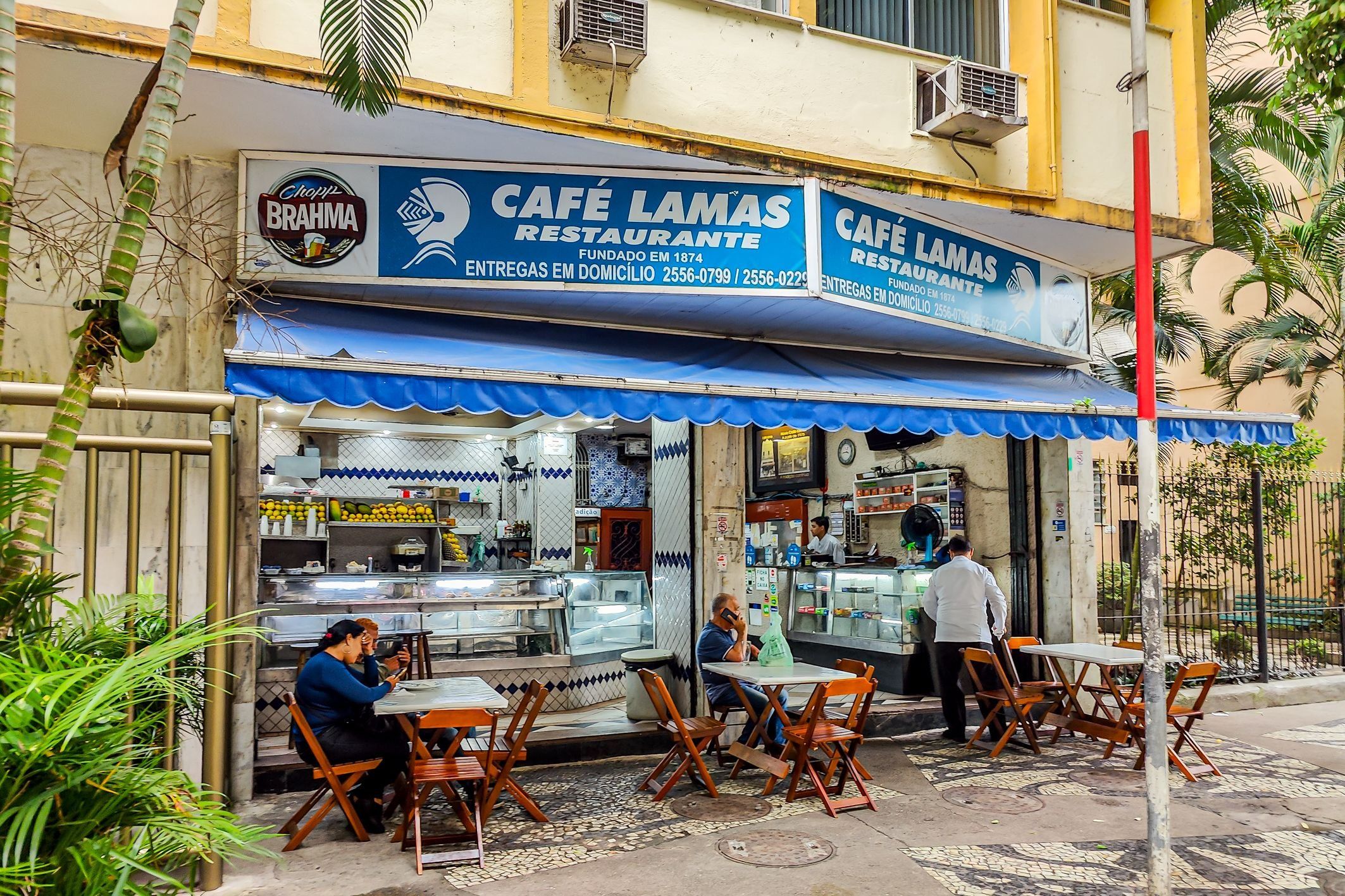 Um café com mesas ao ar livre, toldo azul e placa indicando que é um restaurante.