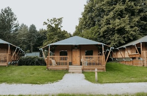 A row of wooden cabins with a porch and a covered entrance, surrounded by trees.