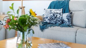 Ein Wohnzimmer mit einem grauen Sofa, einer blau-weißen Decke mit Muster und einem Holztisch mit Blumen in einer Vase.