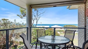 Ein Balkon mit rundem Tisch und Stühlen, der auf eine Küstenlandschaft mit Meer und blauem Himmel blickt.