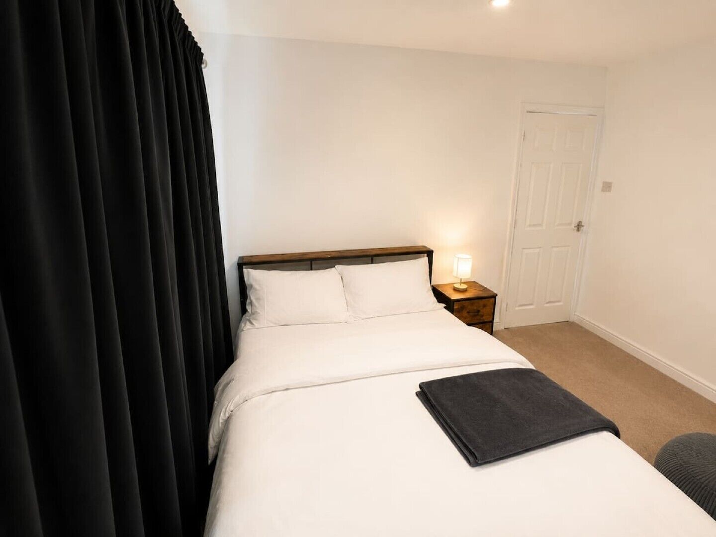 A hotel room with a bed, a nightstand, a lamp, a door, and a curtain.