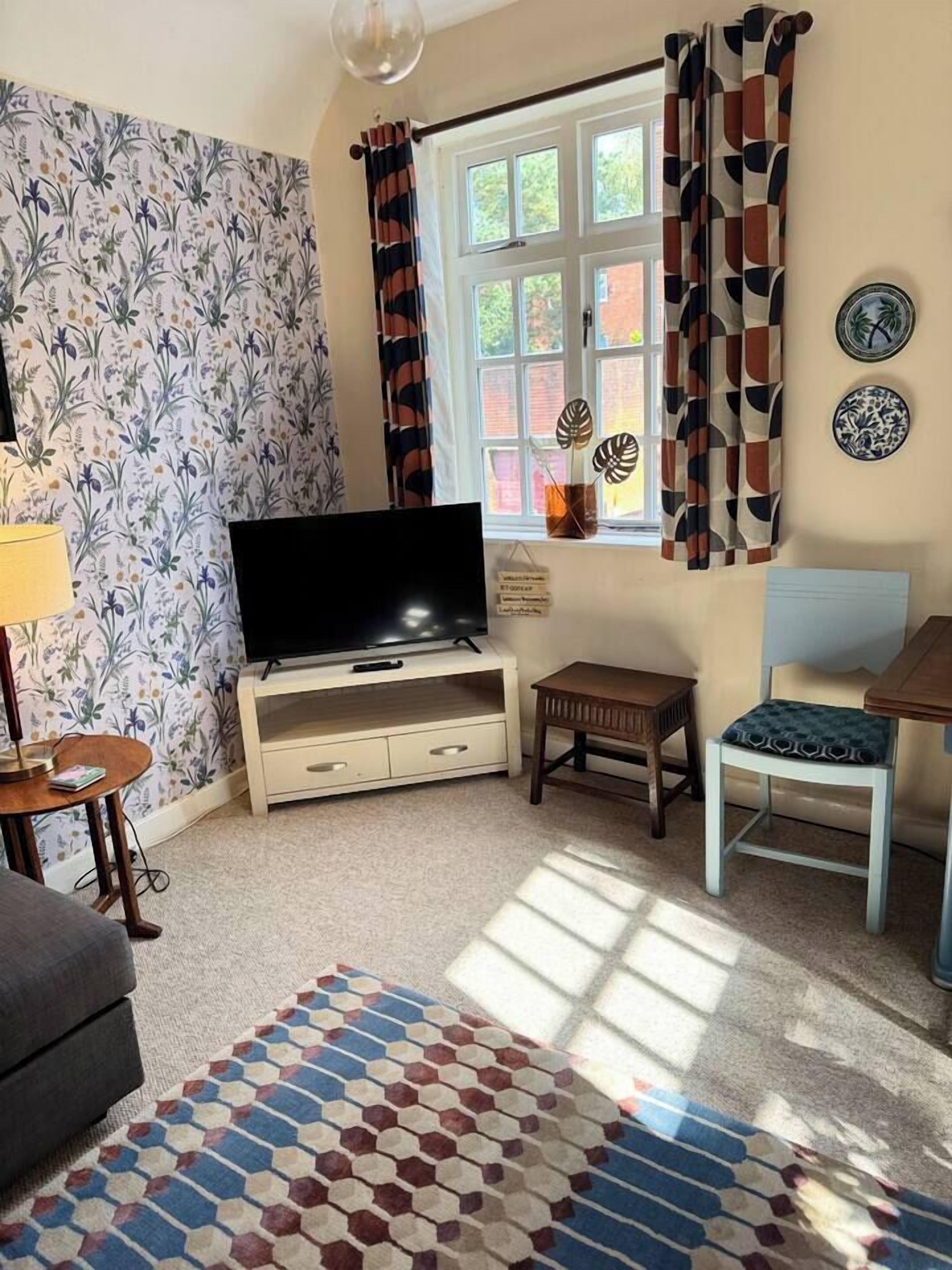 Una sala de estar con televisor de pantalla plana, una mesita, una silla y un tapete estampado.