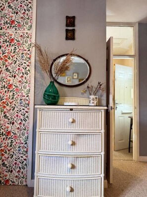 Quarto com penteadeira de vime branco, espelho e papel de parede floral.