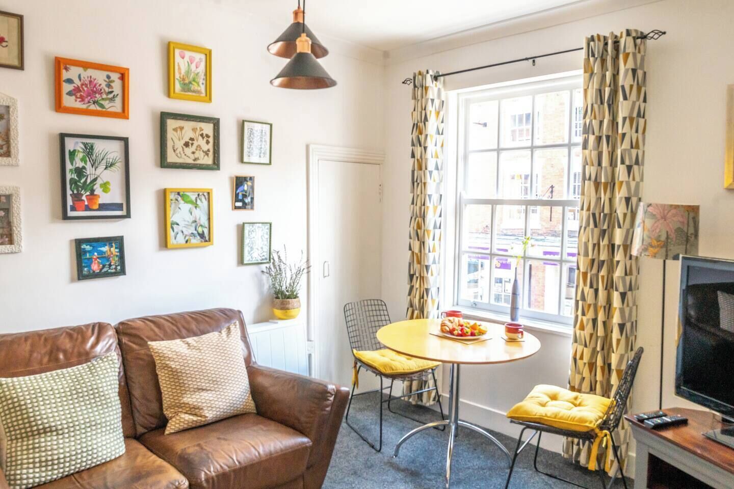 A living room with a brown leather sofa, a round table with chairs, a television, and a window with patterned curtains.