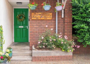 Een groene deur met een krans, een houten bord met tekeningen van vogels, en roze bloemen in een bloempot.