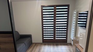 Um quarto com sofá cinzento, piso de madeira, porta deslizante com persianas horizontais e ar condicionado integrado.