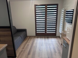 Ein Zimmer mit grauem Sofa, Holzboden, einer Schiebetür mit horizontalen Jalousien und einer eingebauten Klimaanlage.