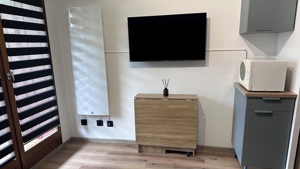 A room with a flat-screen TV mounted on the wall, a wooden cabinet with drawers, a small grey cabinet, and a window with blinds.