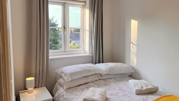 Quarto com cama, mesa de cabeceira, janela com cortinas e vista para o exterior.