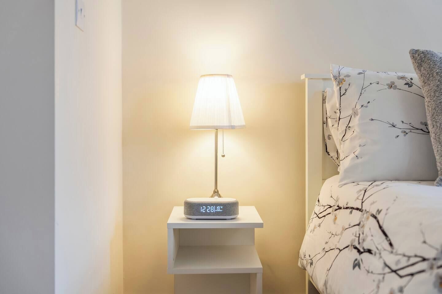 A bedside table with a lamp and a digital clock, a bed with floral bedding, and a wall with a light switch.