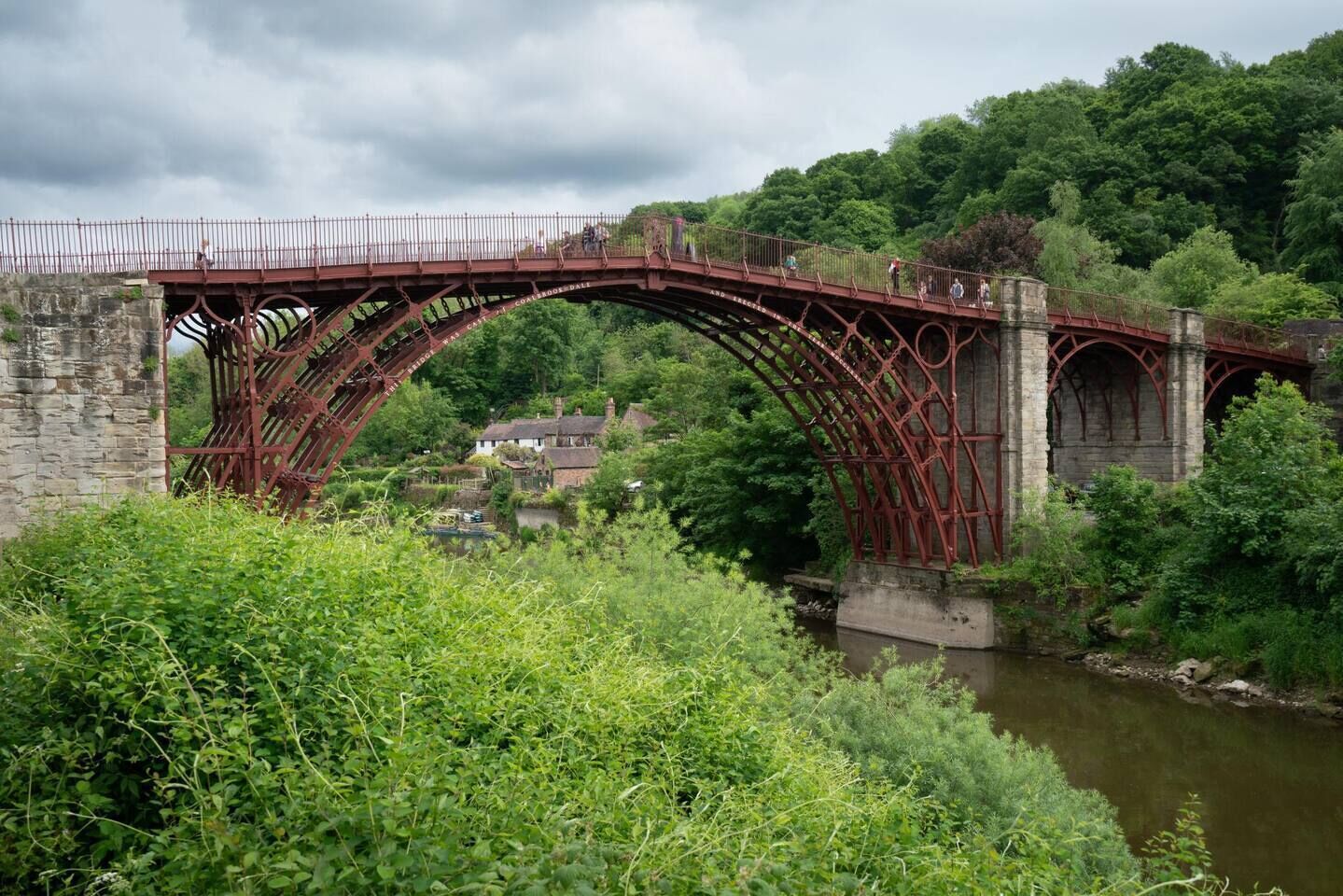Un puente metálico rojo sobre un río, con una casa y vegetación al fondo.