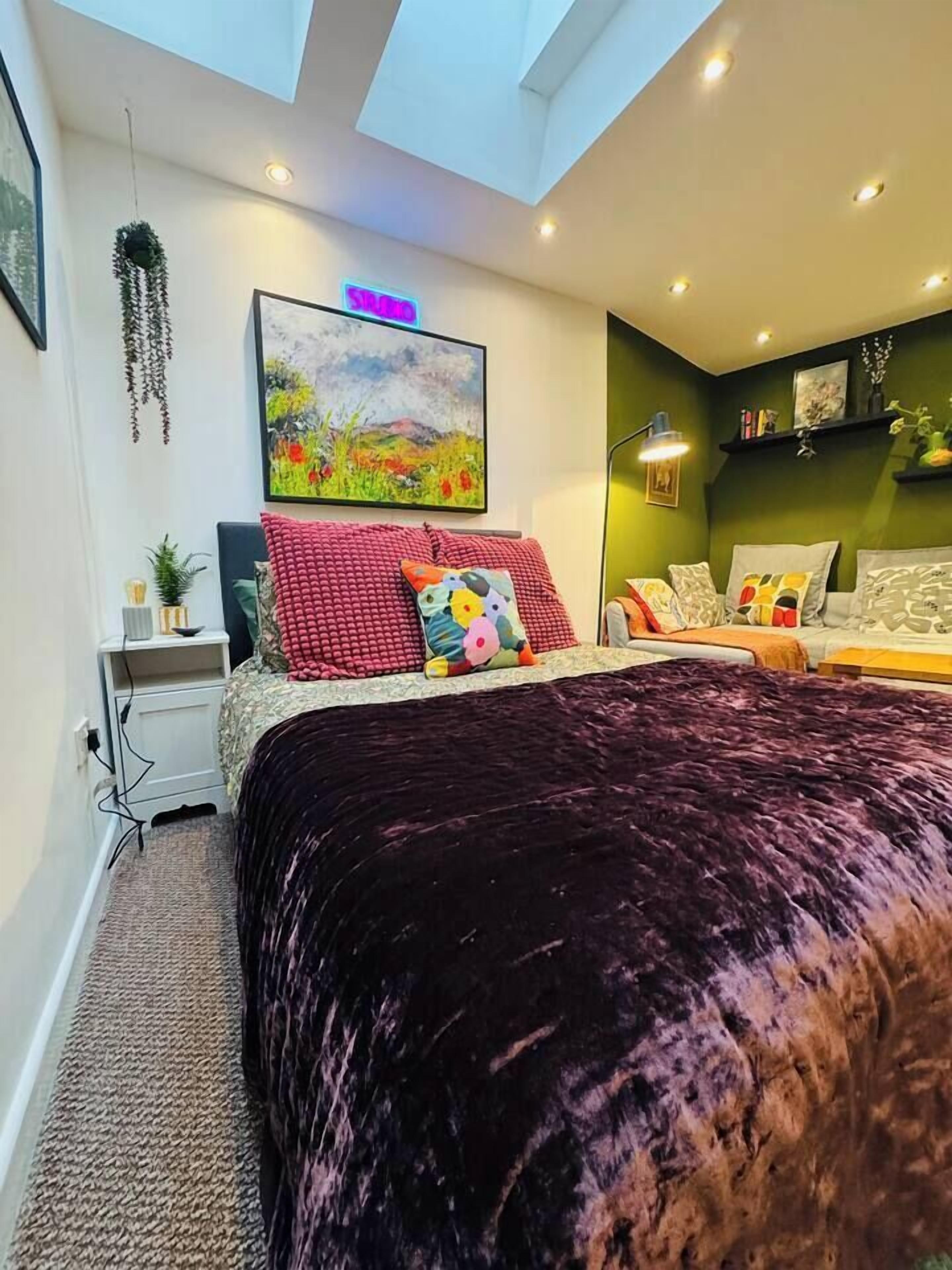 A bedroom with a large bed, a desk, a chair, a hanging plant, and a painting on the wall.
