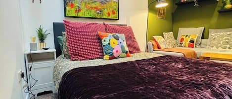 A bedroom with a large bed, a desk, a chair, a hanging plant, and a painting on the wall.