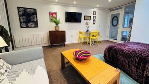 Ein modernes Wohnzimmer mit einer Couch, einem Holztisch mit einer bunten Schüssel, einem Fernseher und einem Essbereich mit Stühlen.