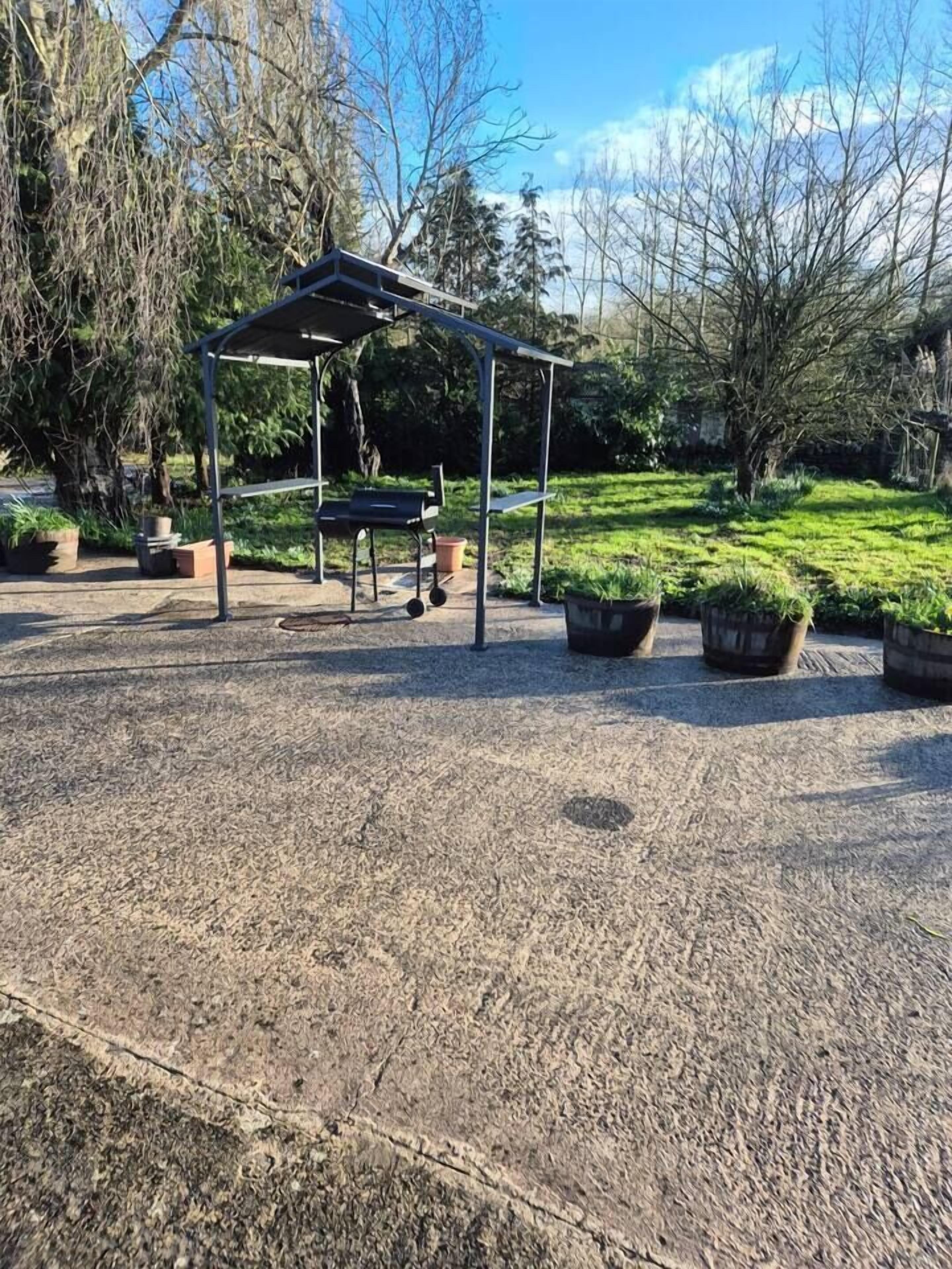 A covered outdoor seating area with a metal roof, surrounded by potted plants and trees.