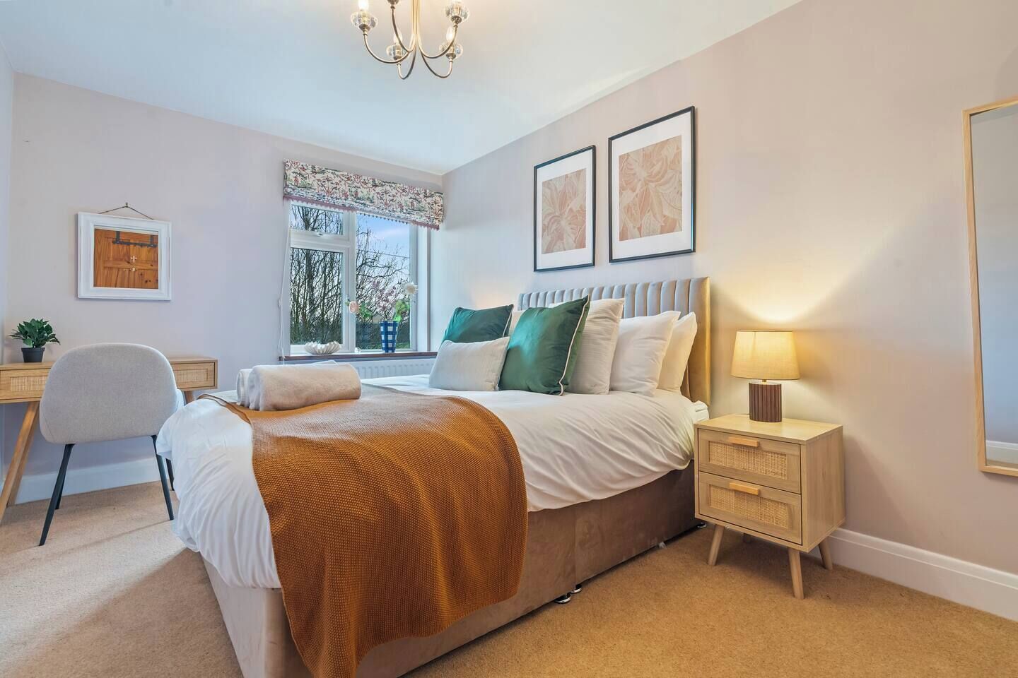 A bedroom with a bed, bedside table, lamp, chair, desk, window with curtains, and framed pictures on the wall.