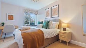 A bedroom with a bed, bedside table, lamp, chair, desk, window with curtains, and framed pictures on the wall.