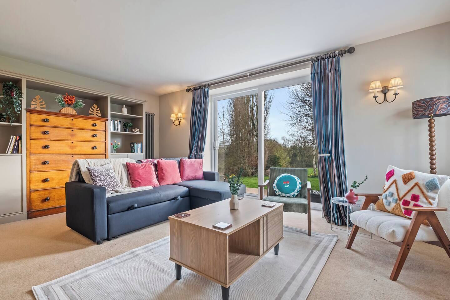 Een moderne woonkamer met een bank, een houten salontafel en een stoel. Er is een boekenplank met decoratieve objecten en een uitzicht op het buitengebied door grote ramen.