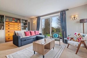 A modern living room with a sofa, a wooden coffee table, and a chair. There is a bookshelf with decorative items and a view of the outdoors through large windows. - #110 Charming Country Stay at Brook Farm, Somerset (Whatley)