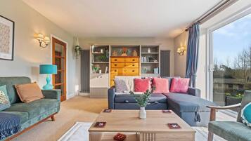 A modern living room with a sofa, armchair, and coffee table. There is a bookshelf with decorative items and a view through a large window.
