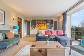 A modern living room with a sofa, armchair, and coffee table. There is a bookshelf with decorative items and a view through a large window. - #110 Charming Country Stay at Brook Farm, Somerset (Whatley)