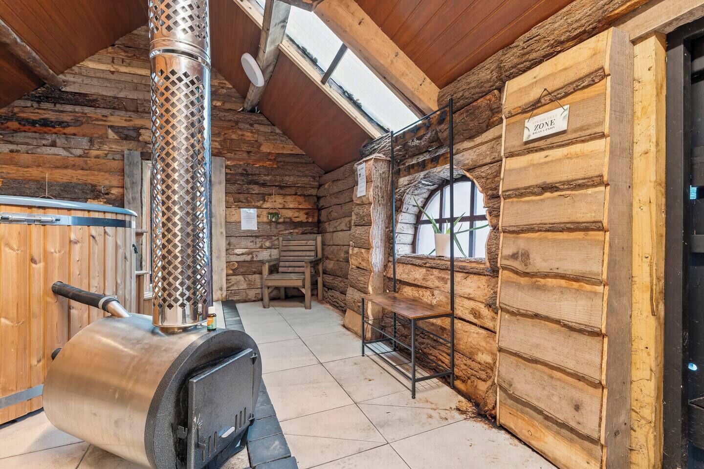 A rustic log cabin interior with a wood-burning stove, a bench, and a window with a view.
