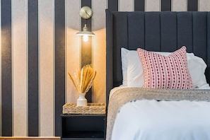 A bed with a dark headboard, white bedding, and a patterned pillow. A bedside table with a woven basket and a vase of dried plants is visible.