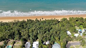 An aerial view of a coastal area with a beach, dense forest, and residential buildings with solar panels.