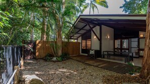 A covered outdoor seating area with a wooden deck, surrounded by tropical vegetation and a wooden fence.