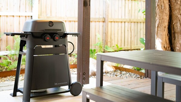 A portable grill with a digital display and control knobs, positioned on a patio with a wooden fence and greenery in the background.