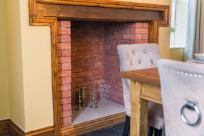 A fireplace with a brick interior, a wooden mantel, and a table with a chair in front.