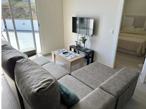 Ein modernes Wohnzimmer mit einer Couch, einem Couchtisch, einem Fernseher und einem Bett im Hintergrund.