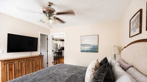 Una camera da letto con un letto, una ventola a soffitto, una TV, mobili in legno e una cucina sullo sfondo.
