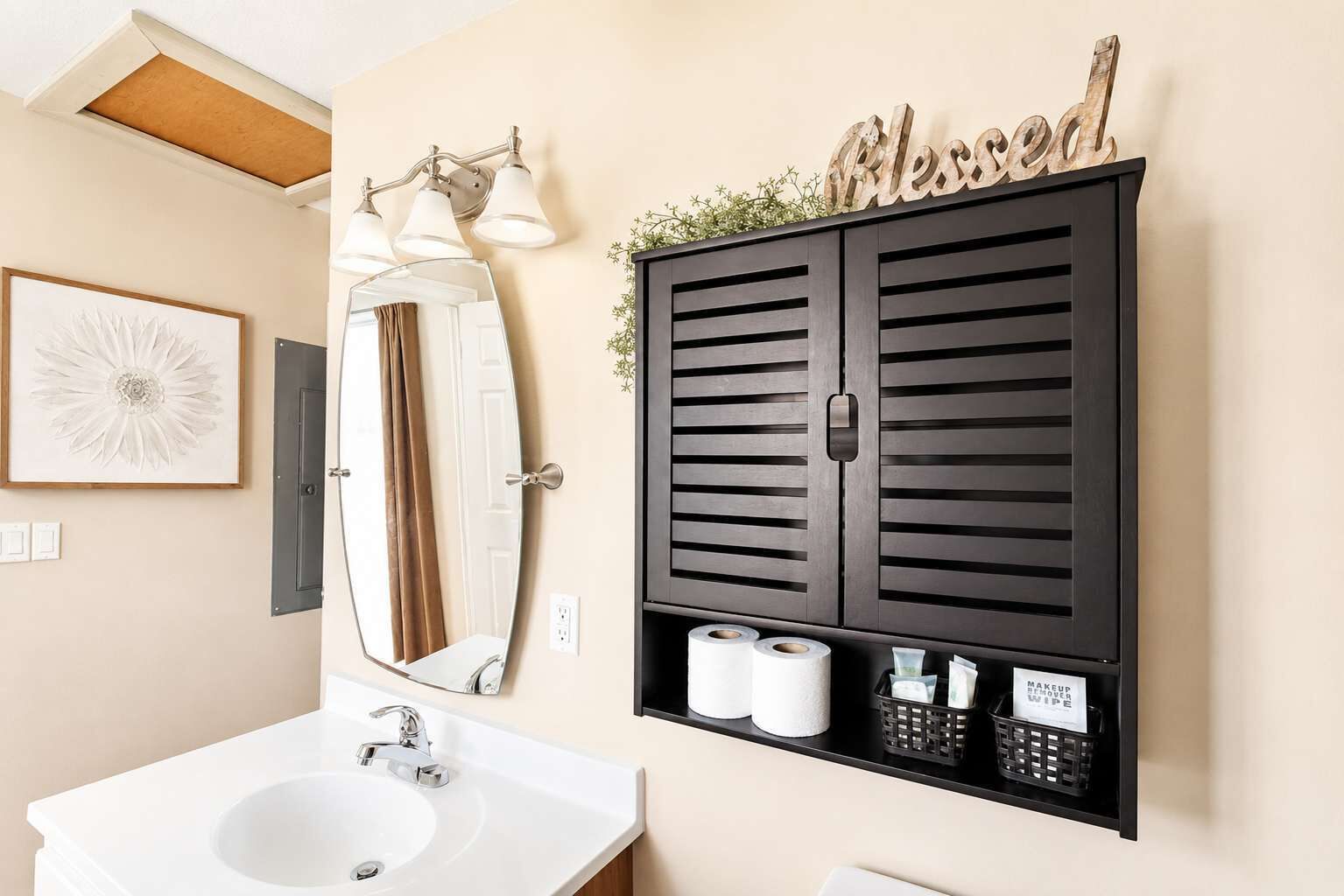 A bathroom with a white sink, a large mirror, a black cabinet with a 'Blessed' sign, and a framed artwork on the wall.
