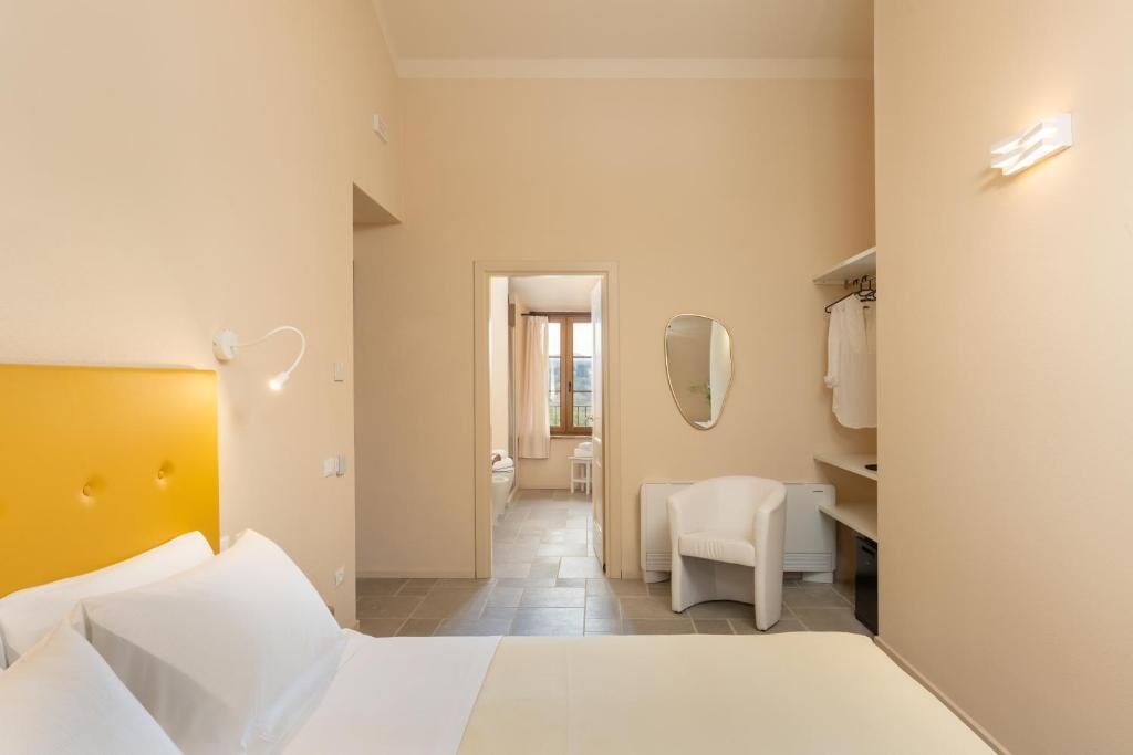 A hotel room with a bed, a bedside table, a chair, a mirror, and a small bathroom area.