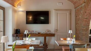 Un área de comedor con pared de ladrillo, un televisor de pantalla plana sobre una mesa de madera, y una puerta a la derecha.