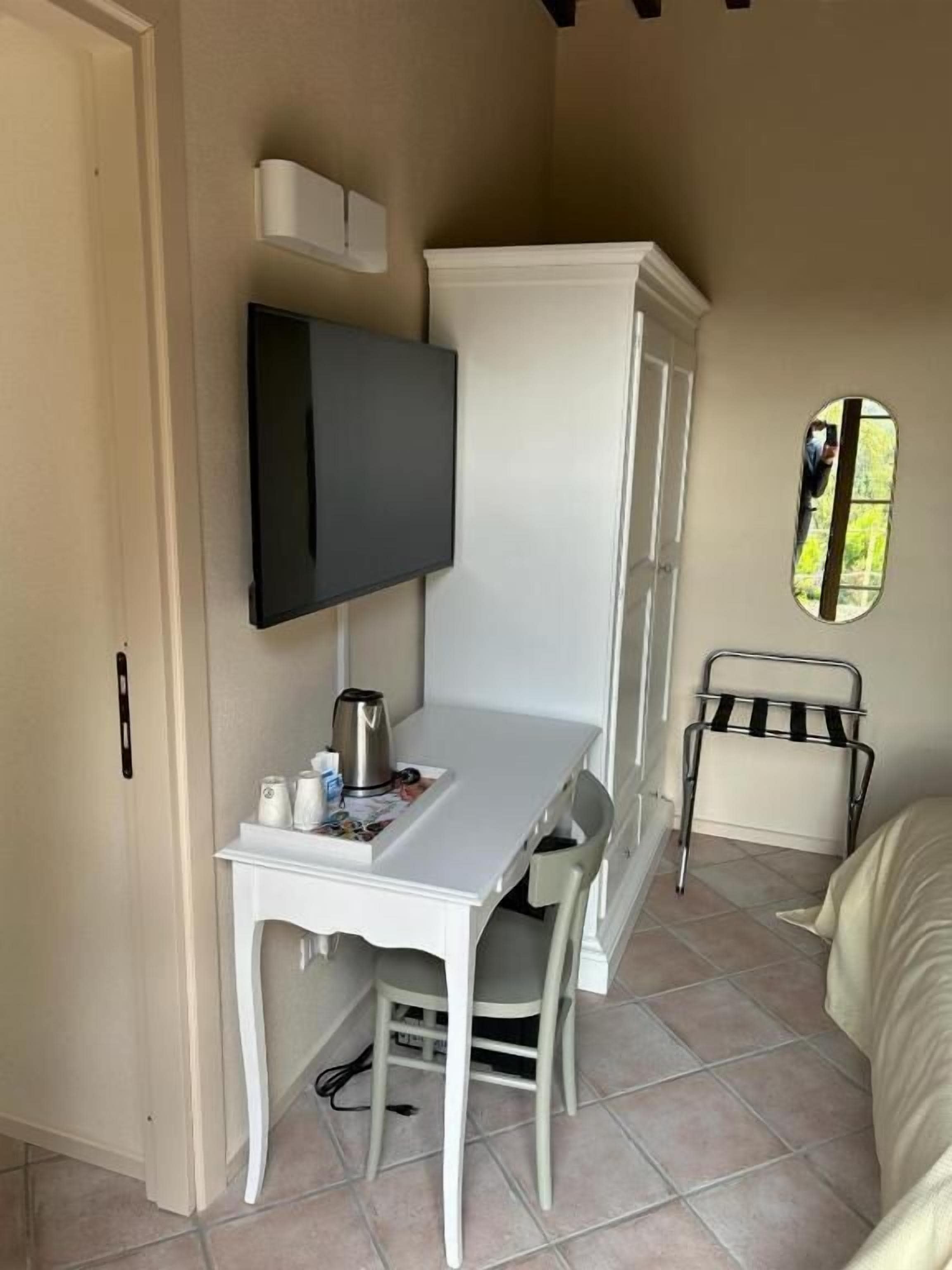 Ein kleines Zimmer mit einem weißen Schreibtisch, einem Stuhl, einem an der Wand befestigten Fernseher, einem kleinen Fenster und einem Metallregal.
