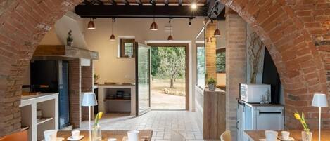 A dining area with a brick archway, wooden tables, and chairs, connected to a kitchen and a view of the outdoors.
