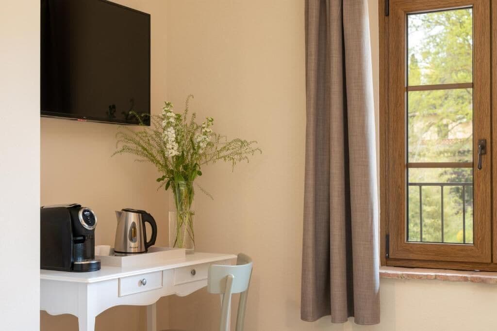 Ein Zimmer mit einem weißen Schreibtisch, einer Kaffeemaschine, einer Vase mit Blumen, einem über dem Schreibtisch montierten Fernseher und einem Fenster mit Vorhängen.