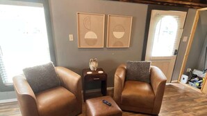 Una habitación con dos sillones de cuero, un pequeño reposapiés y una mesita de noche con un reloj. Dos obras de arte abstracto enmarcadas decoran la pared sobre el reposapiés.